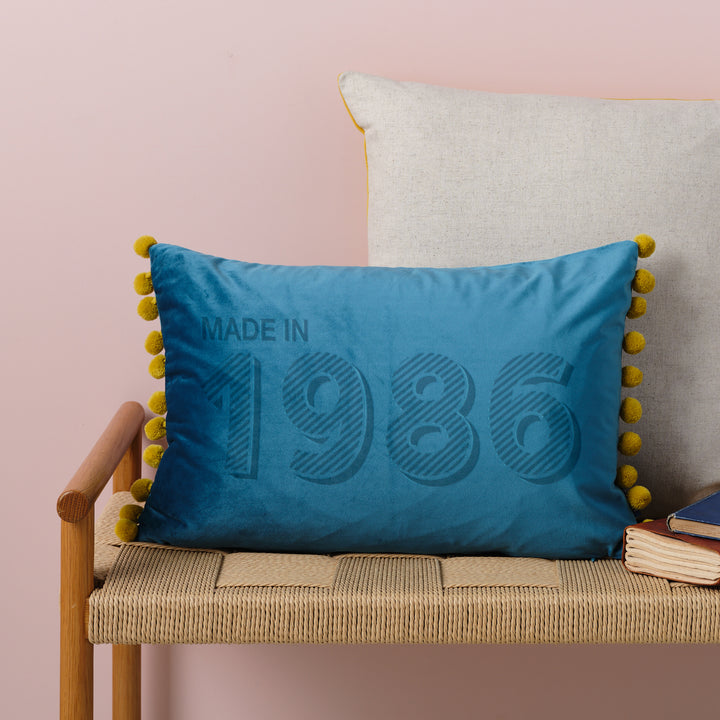 A personalised 40th birthday gift velvet cushion with 'MADE IN 1986' laser engraved on it, placed on a wicker bench with other cushions and a book.