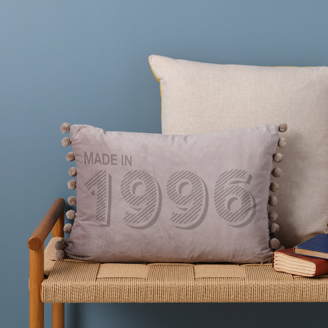 A personalised 30th birthday gift velvet cushion with 'MADE IN 1996' laser engraved on it, placed on a wicker bench with other cushions and a book.