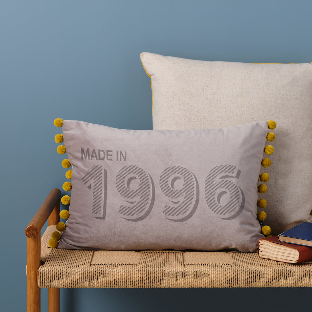 A personalised 30th birthday gift velvet cushion with 'MADE IN 1996' laser engraved on it, placed on a wicker bench with other cushions and a book.