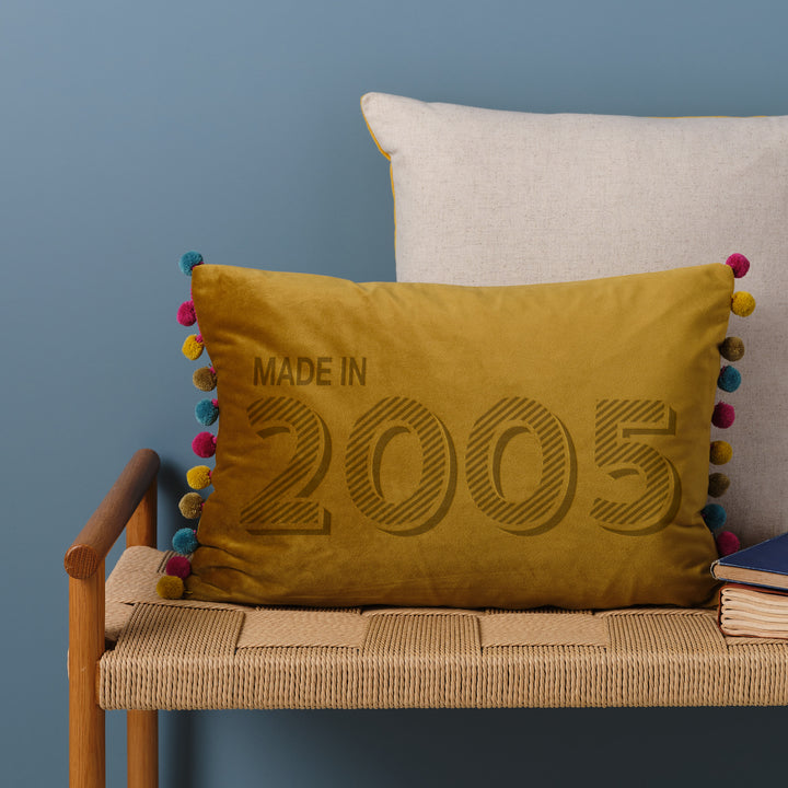 A personalised 21st birthday gift velvet cushion with 'MADE IN 2005' laser engraved on it, placed on a wicker bench with other cushions and a book.