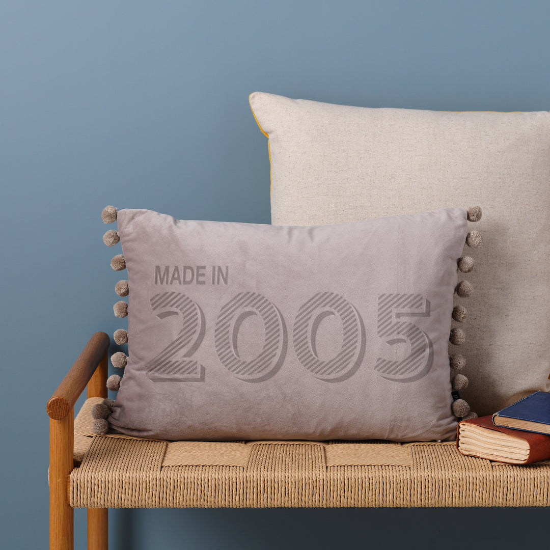 A personalised 21st birthday gift velvet cushion with 'MADE IN 2005' laser engraved on it, placed on a wicker bench with other cushions and a book.