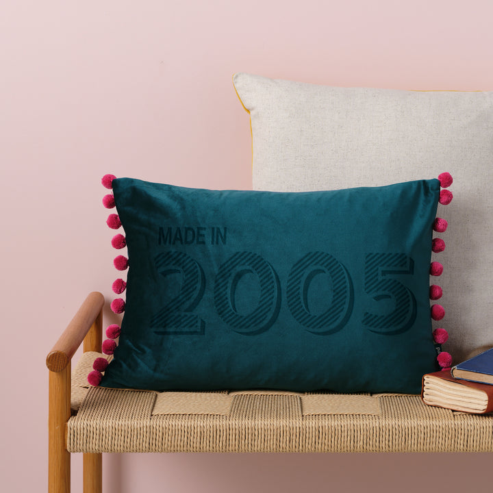 A personalised 21st birthday gift velvet cushion with 'MADE IN 2005' laser engraved on it, placed on a wicker bench with other cushions and a book.