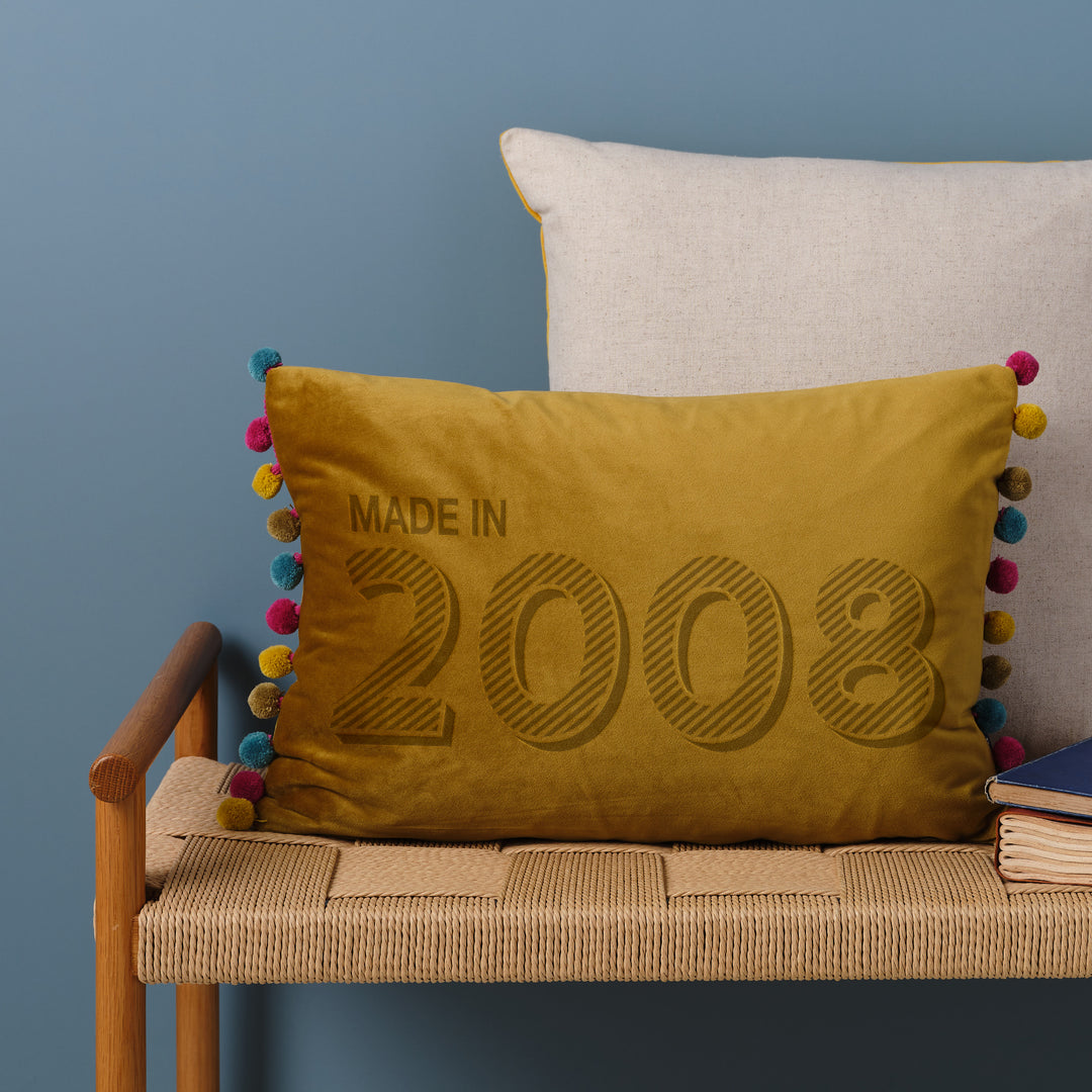 A personalised 18th birthday gift velvet cushion with 'MADE IN 2008' laser engraved on it, placed on a wicker bench with other cushions and a book.