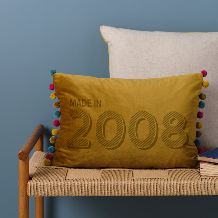 A personalised 18th birthday gift velvet cushion with 'MADE IN 2008' laser engraved on it, placed on a wicker bench with other cushions and a book.