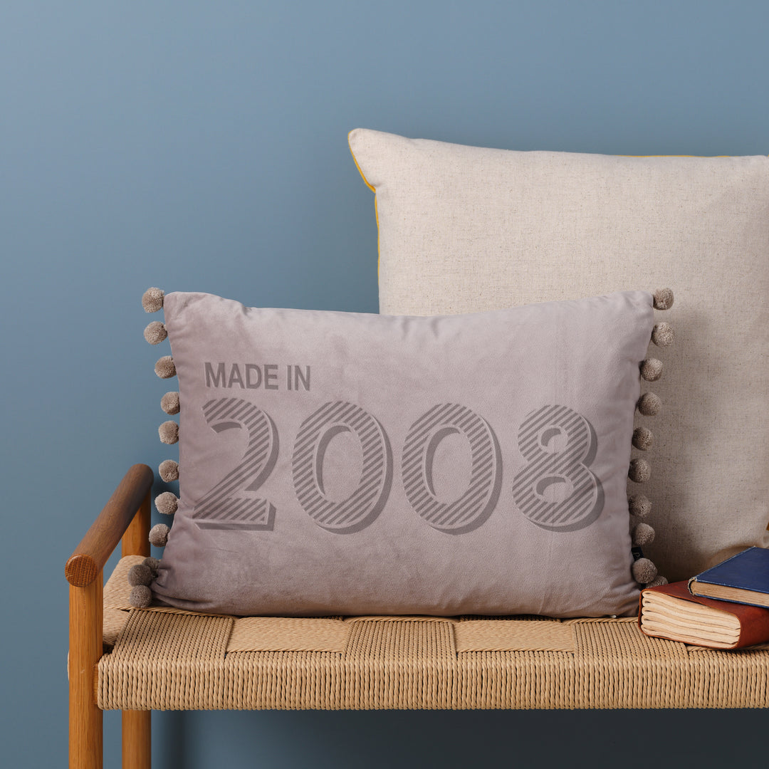 A personalised 18th birthday gift velvet cushion with 'MADE IN 2008' laser engraved on it, placed on a wicker bench with other cushions and a book.