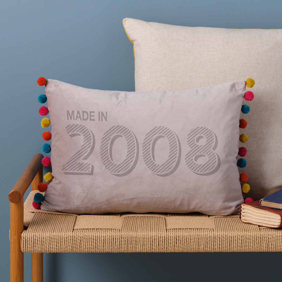 A personalised 18th birthday gift velvet cushion with 'MADE IN 2008' laser engraved on it, placed on a wicker bench with other cushions and a book.