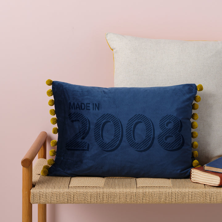 A personalised 18th birthday gift velvet cushion with 'MADE IN 2008' laser engraved on it, placed on a wicker bench with other cushions and a book.