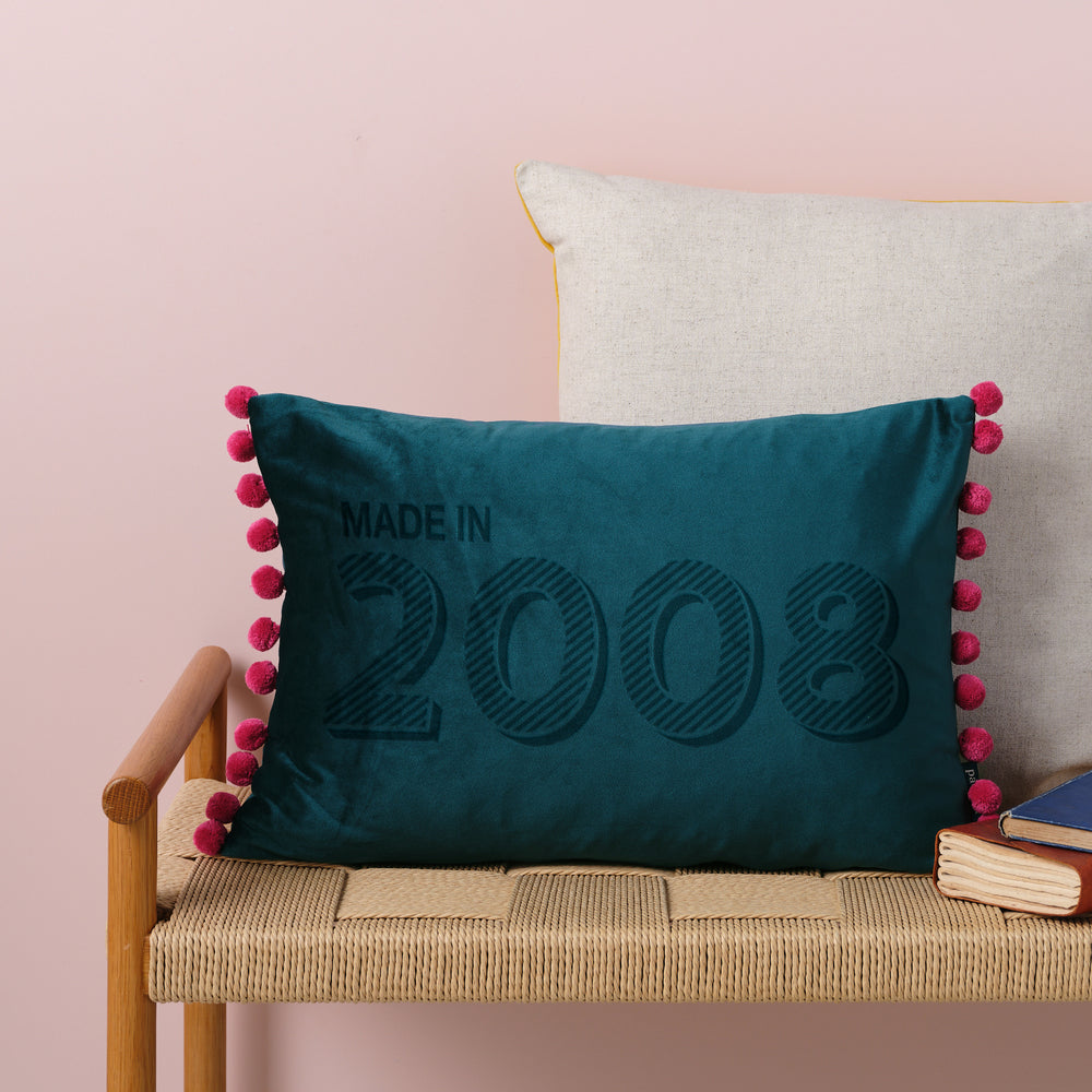 A personalised 18th birthday gift velvet cushion with 'MADE IN 2008' laser engraved on it, placed on a wicker bench with other cushions and a book.