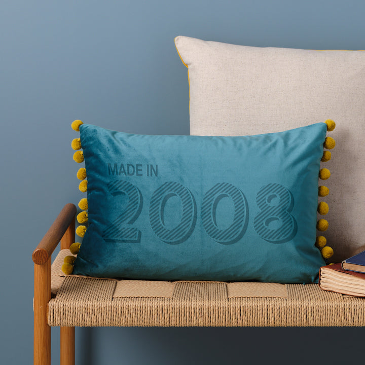 A personalised 18th birthday gift velvet cushion with 'MADE IN 2008' laser engraved on it, placed on a wicker bench with other cushions and a book.