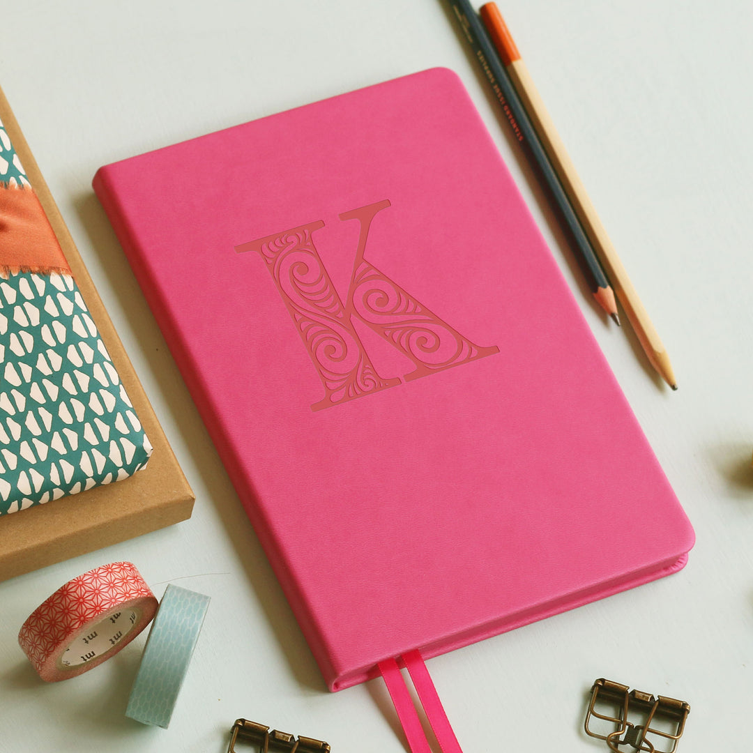 A top down image of a hot pink vegan leather notebook resting on a pale aqua table. The front of the notebook is personalised with the letter K engraved in a large decorative font. Surrounding the book are various stationary items.