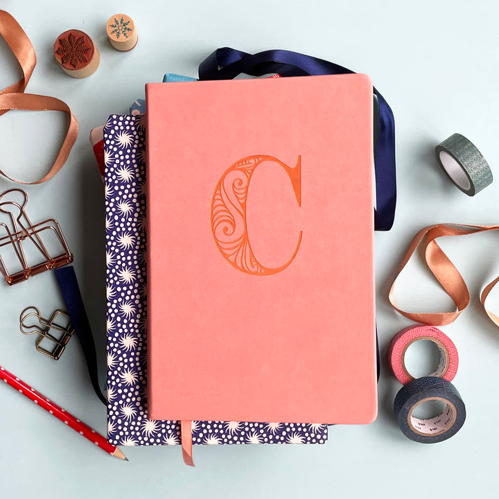 A top down image of a pink vegan leather notebook resting on a pale aqua table. The front of the notebook is personalised with the letter C engraved in a large decorative font. Surrounding the book are various stationary items.