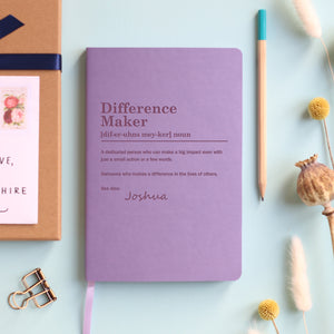 A top down image of a lilac vegan leather notebook resting on a pale aqua table. The front of the notebook is engraved with a bespoke dictionary definition of the phrase Difference Maker and personalised with the name Joshua. Surrounding the book are various stationary items and dried flowers.