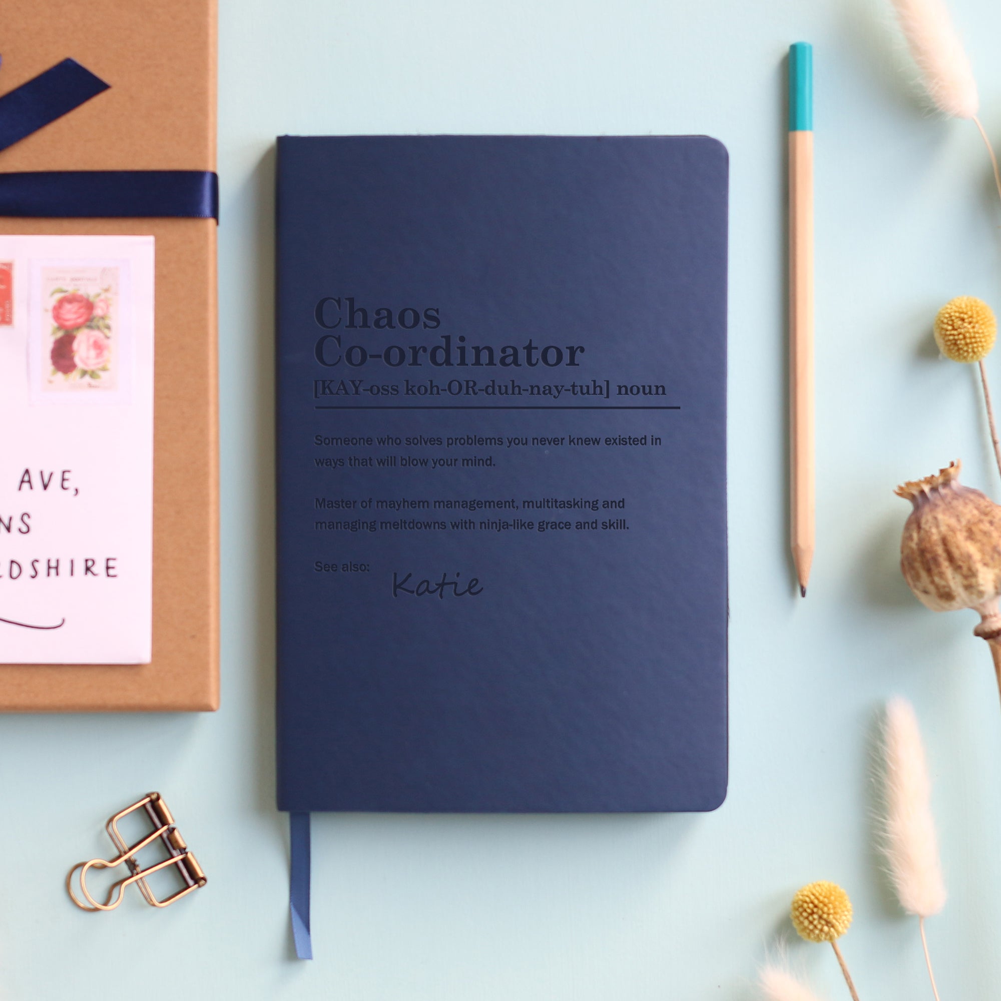 A top down image of a navy blue vegan leather notebook resting on a pale aqua table. The front of the notebook is engraved with a bespoke dictionary definition of the phrase Chaos-Co-ordinator and personalised with the name Katie. Surrounding the book are various stationary items and dried flowers.