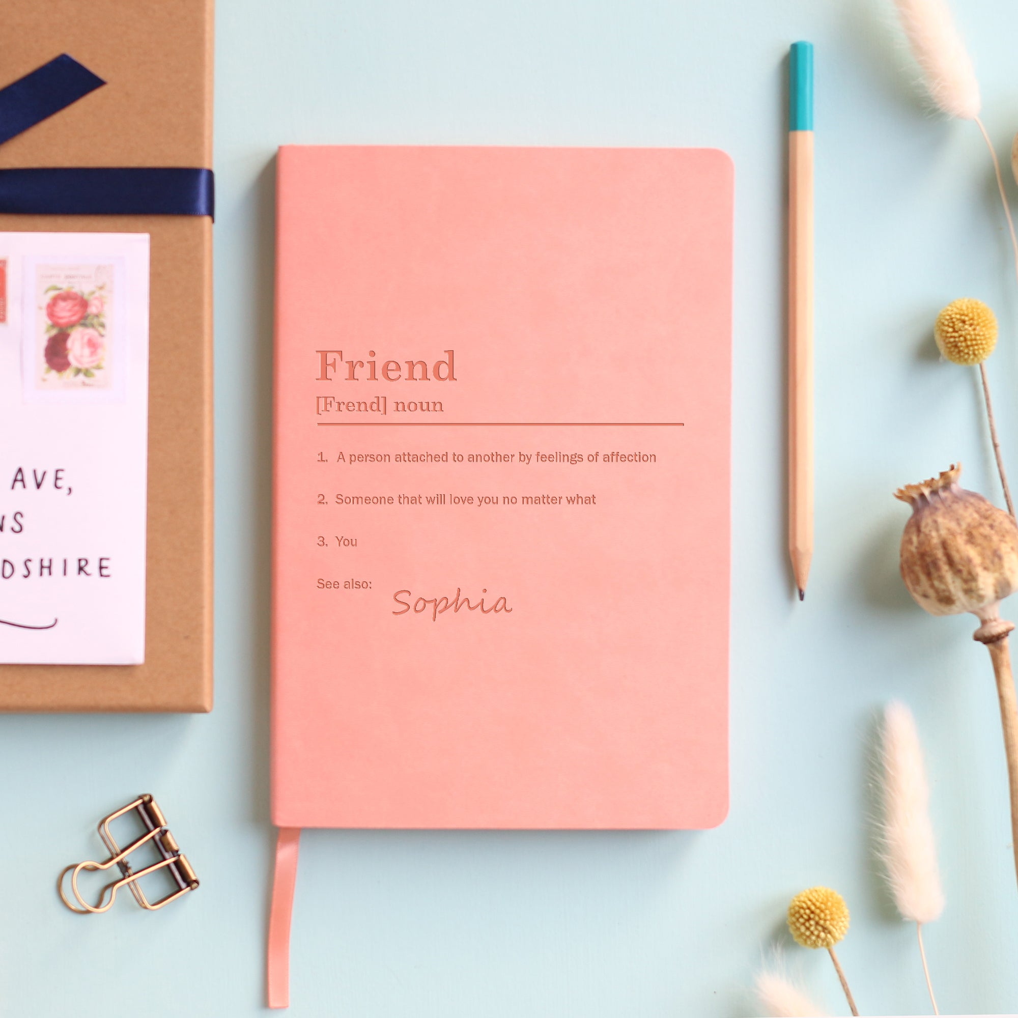 A top down image of a blush pink vegan leather notebook resting on a pale aqua table. The front of the notebook is engraved with a bespoke dictionary definition of the word Friend and personalised with the name Sophia. Surrounding the book are various stationary items and dried flowers.