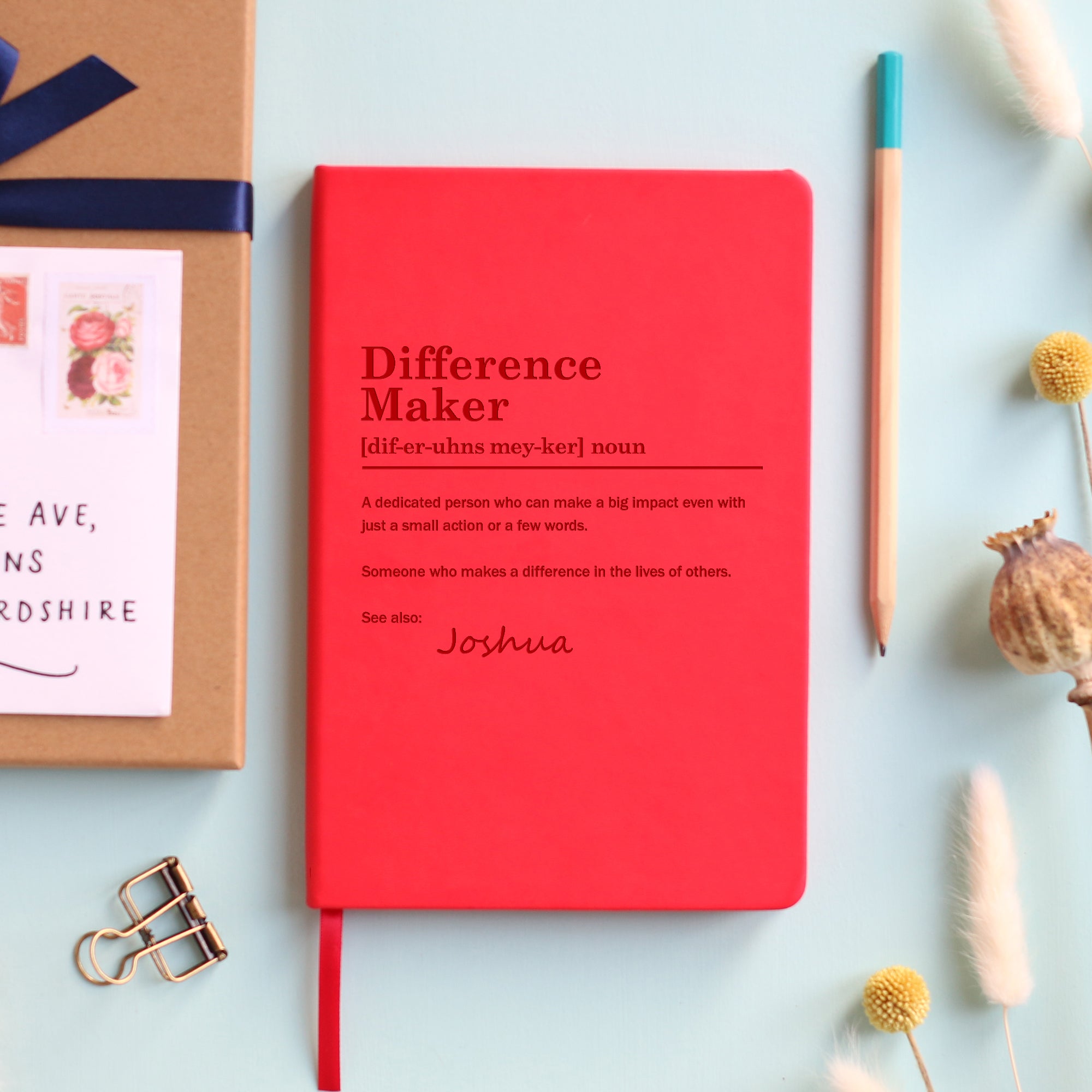 A top down image of a red vegan leather notebook resting on a pale aqua table. The front of the notebook is engraved with a bespoke dictionary definition of the phrase Difference Maker and personalised with the name Joshua. Surrounding the book are various stationary items and dried flowers.