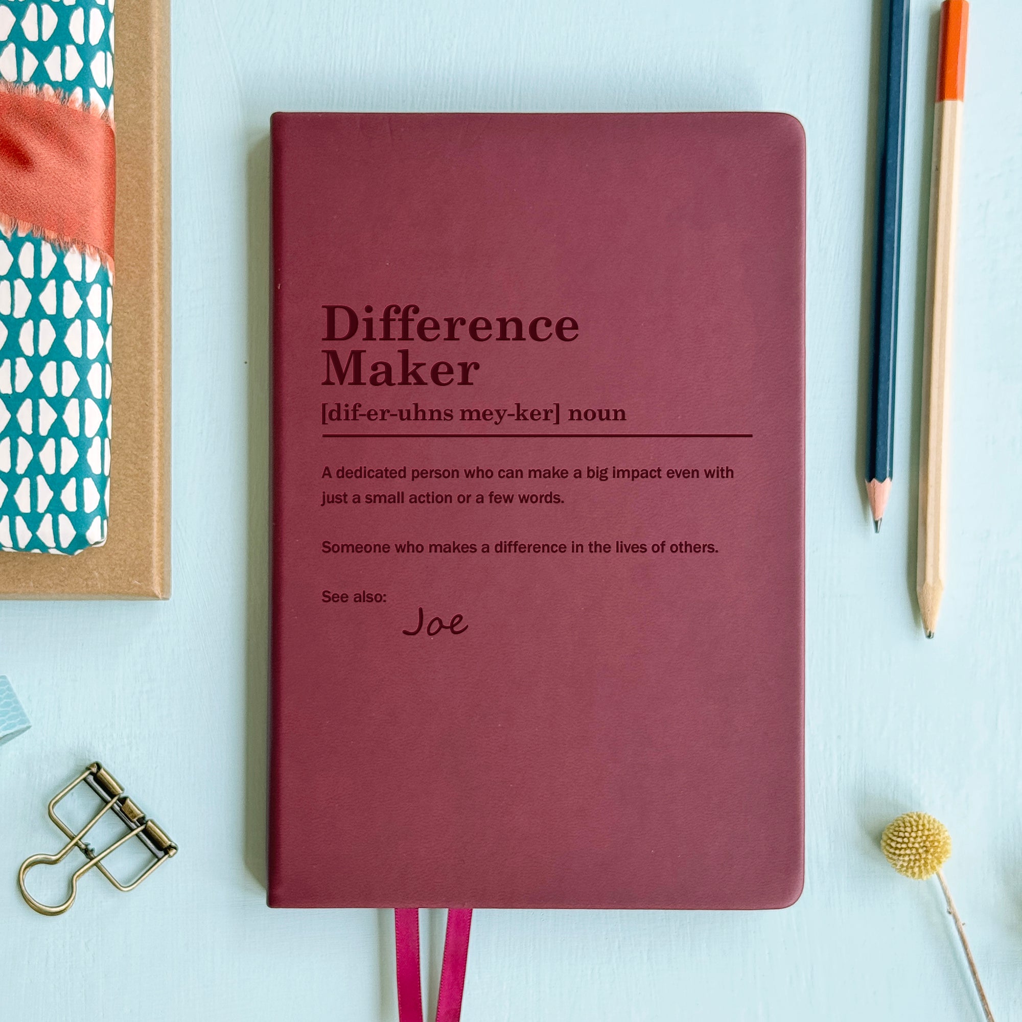 A top down image of a burgundy vegan leather notebook resting on a pale aqua table. The front of the notebook is engraved with a bespoke dictionary definition of the phrase Difference Maker and personalised with the name Joe. Surrounding the book are various stationary items.