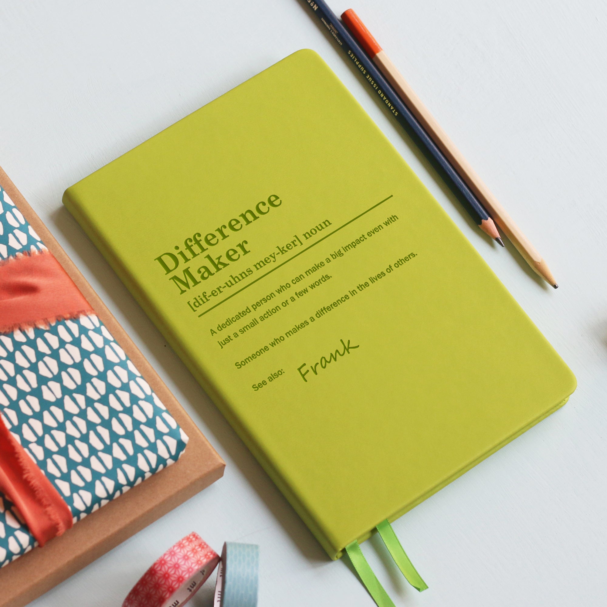 A top down image of a lime green vegan leather notebook resting on a pale aqua table. The front of the notebook is engraved with a bespoke dictionary definition of the phrase Difference Maker and personalised with the name Frank. Surrounding the book are various stationary items.