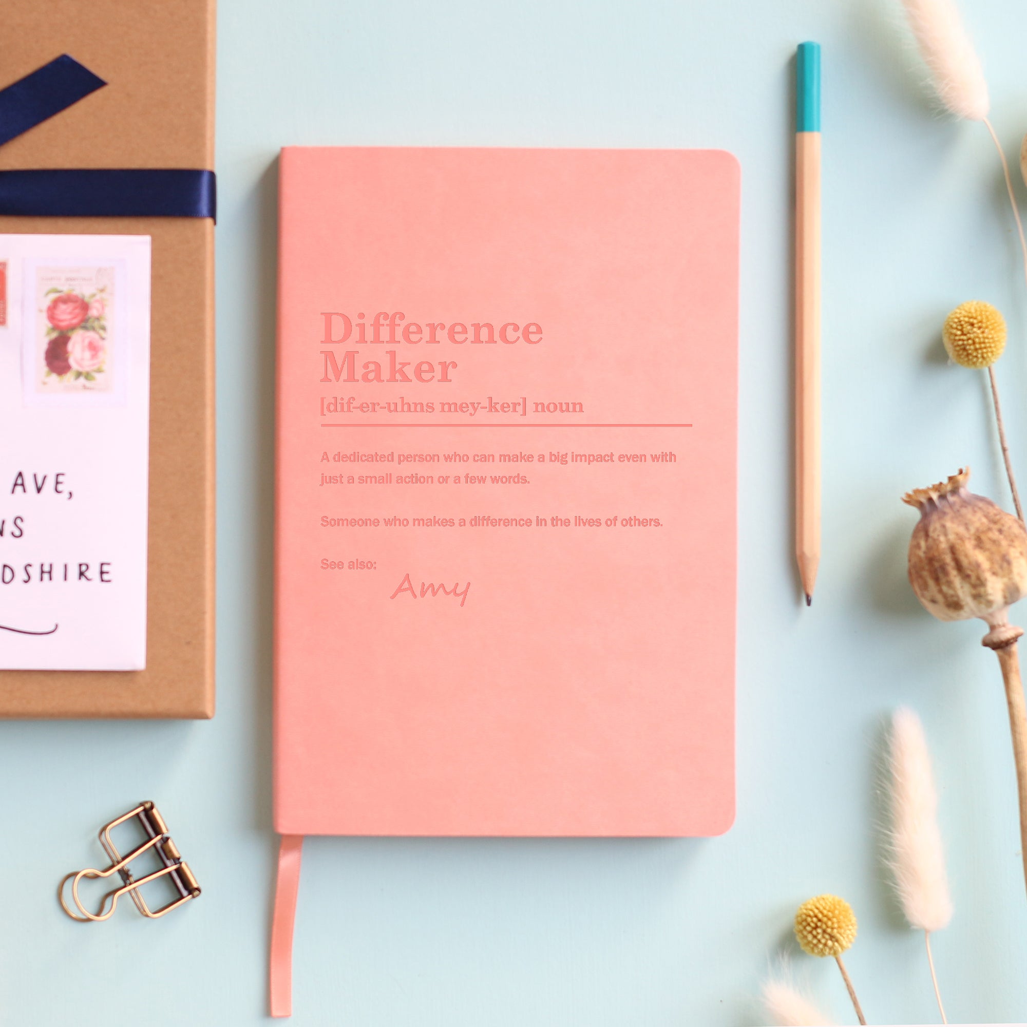 A top down image of a pink vegan leather notebook resting on a pale aqua table. The front of the notebook is engraved with a bespoke dictionary definition of the phrase Difference Maker and personalised with the name Amy. Surrounding the book are various stationary items.