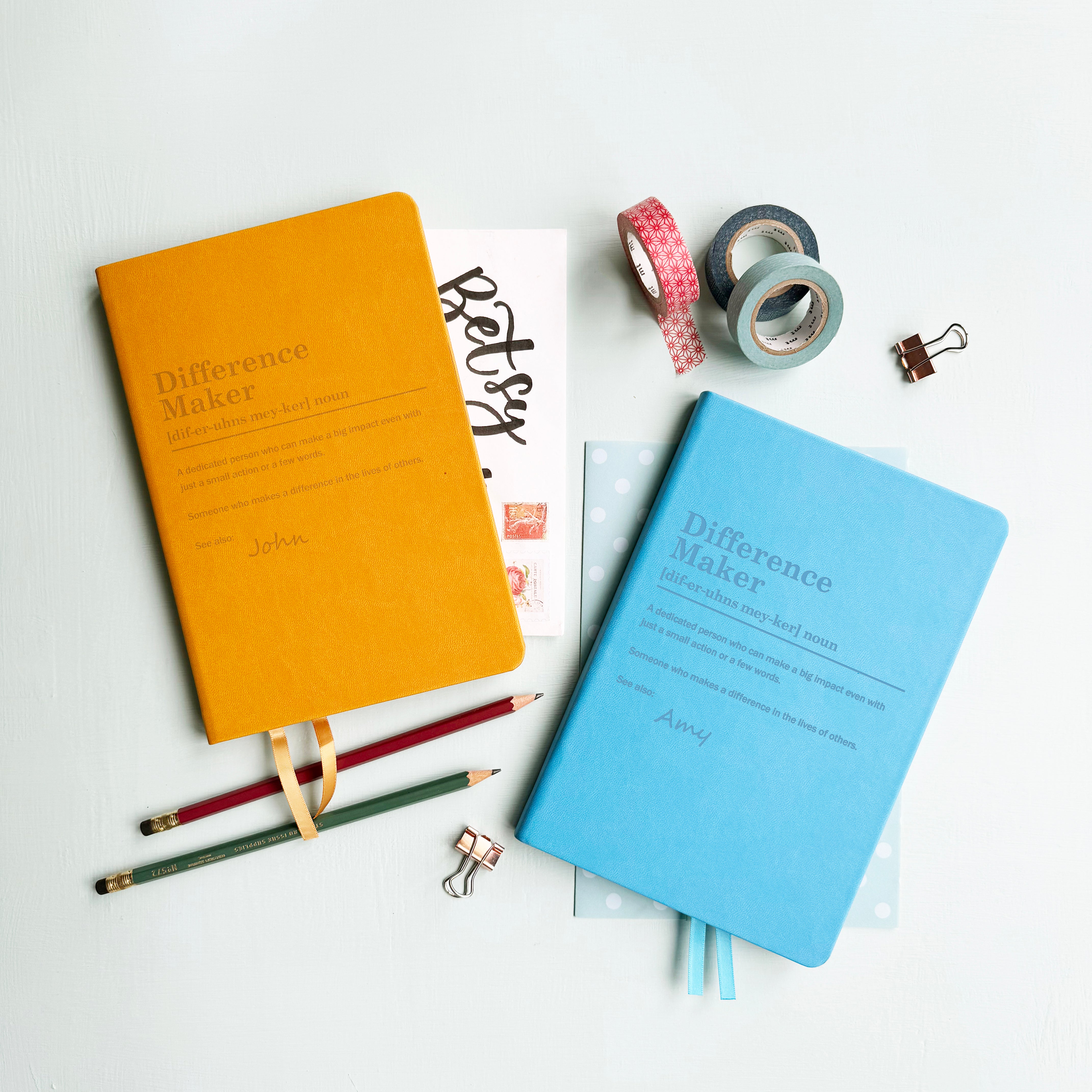 A top down image of two vegan leather notebooks, one mustard yellow and one blue. They are resting on a pale aqua table. The front of each notebook features the words the words Difference Maker and definition text. The name John is engraved on the mustard yellow notebook and Amy is engraved on the blue journal.  Surrounding the book are various stationary items.