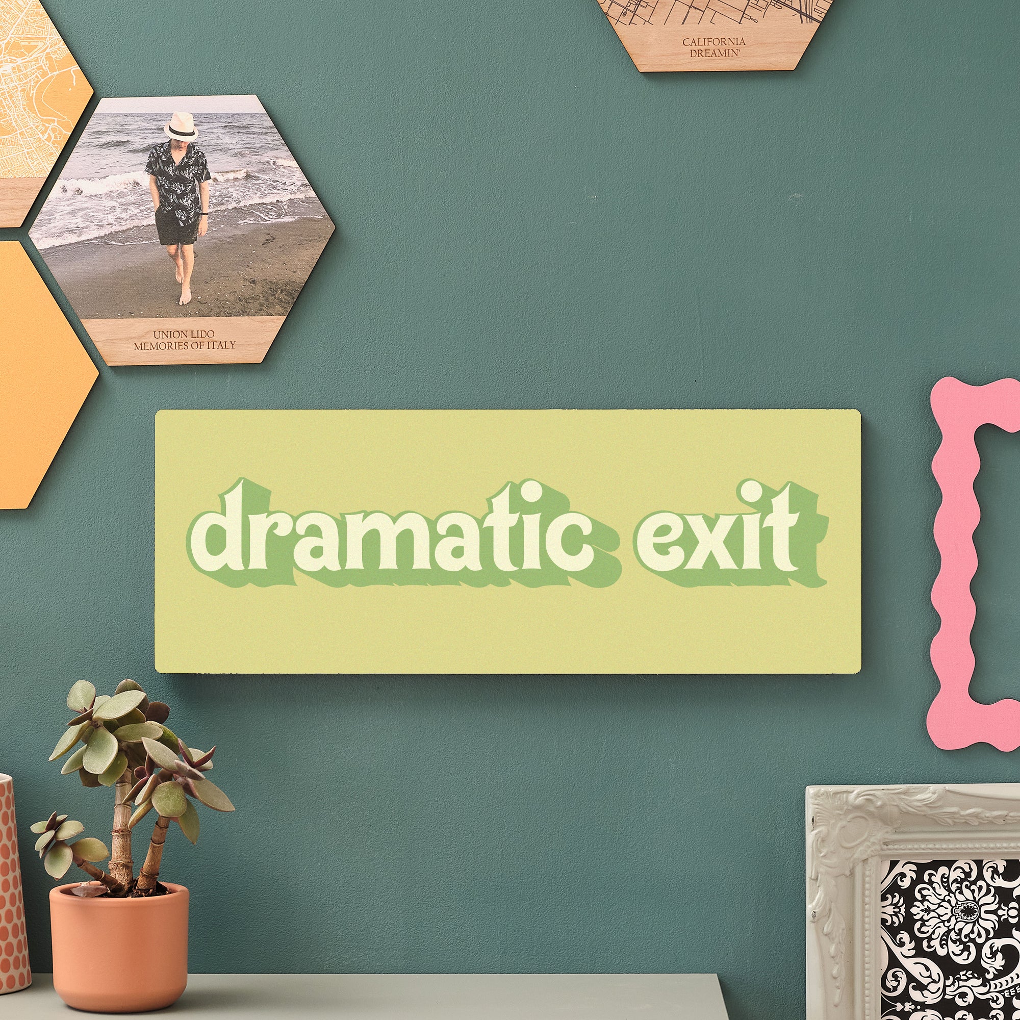 Close-up of a green wooden sign reading “dramatic exit” in playful retro lettering, mounted on a green wall surrounded by hexagon wall tiles and potted plants.