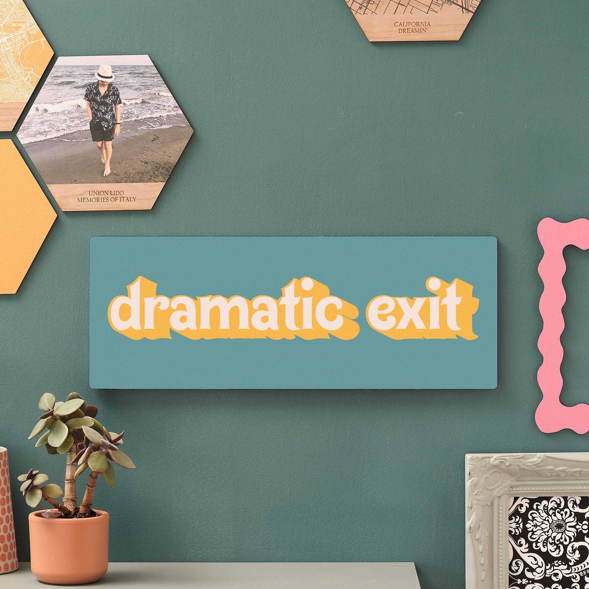 Bold wooden wall sign reading “dramatic exit” in cream and mustard yellow on a teal background, displayed as part of a stylish gallery wall with hexagonal tiles and succulent plants.