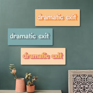 Three wooden “dramatic exit” signs in different colourways—oranges, teals, and a colourway that reminds me of the old fruit salad penny sweets —arranged vertically on a green wall with complementary wall art and home decor elements.
