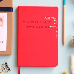 A top down image of a red vegan leather resting on a pale aqua table. The front of the notebook is personalised with the name Mr Williams and engraved with the phrase Evil Genius. Surrounding the book are various stationary items and dried flowers.The image is edited to show the text that can be personalised.