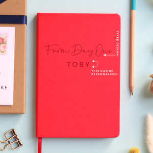 A top down image of a red vegan leather notebook resting on a pale aqua table. The front of the notebook features the phrase "From Day One". It is personalised with the name Toby. Surrounding the book are various stationary items and dried flowers. The image is edited to highlight the area that can be personalised.