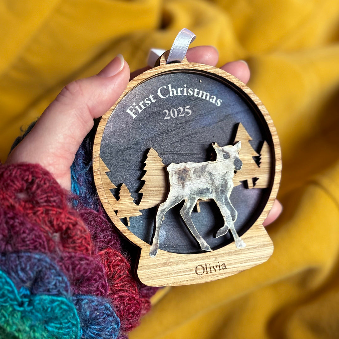 Wooden ornament with a deer silhouette, 'First Christmas 2025', and 'Olivia' text, held against a yellow fabric background.