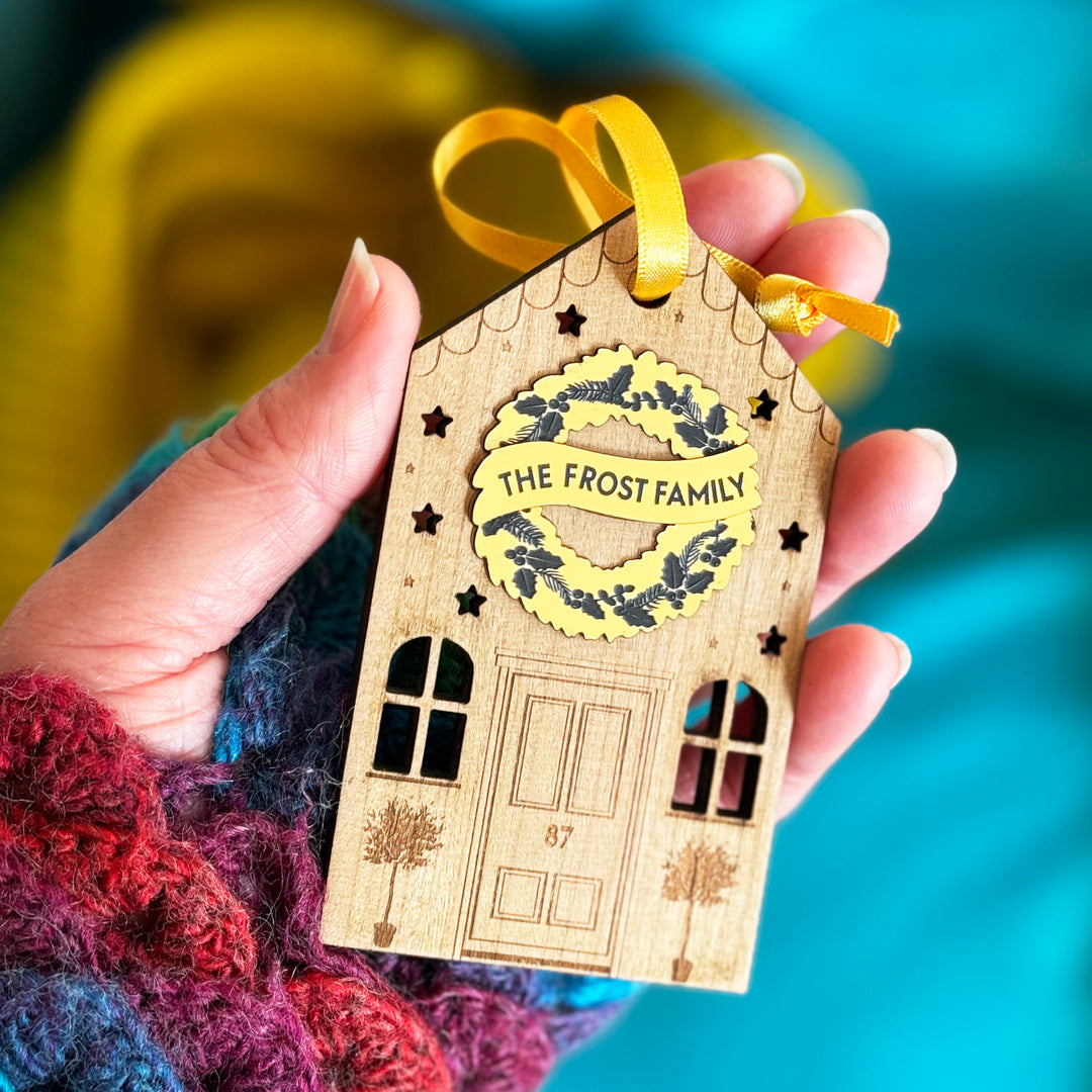 Wooden house Christmas ornament with personalised golden wreath and engraved details being held in a hand.