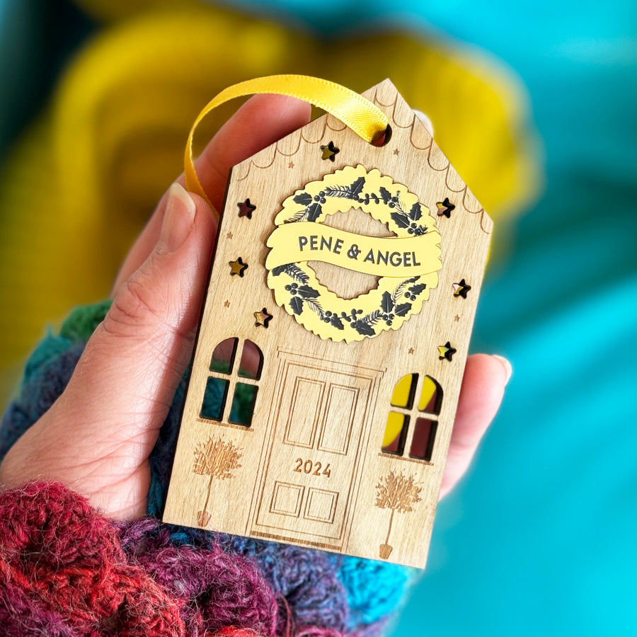 Wooden house Christmas ornament with personalised golden wreath and engraved details being held in a hand.
