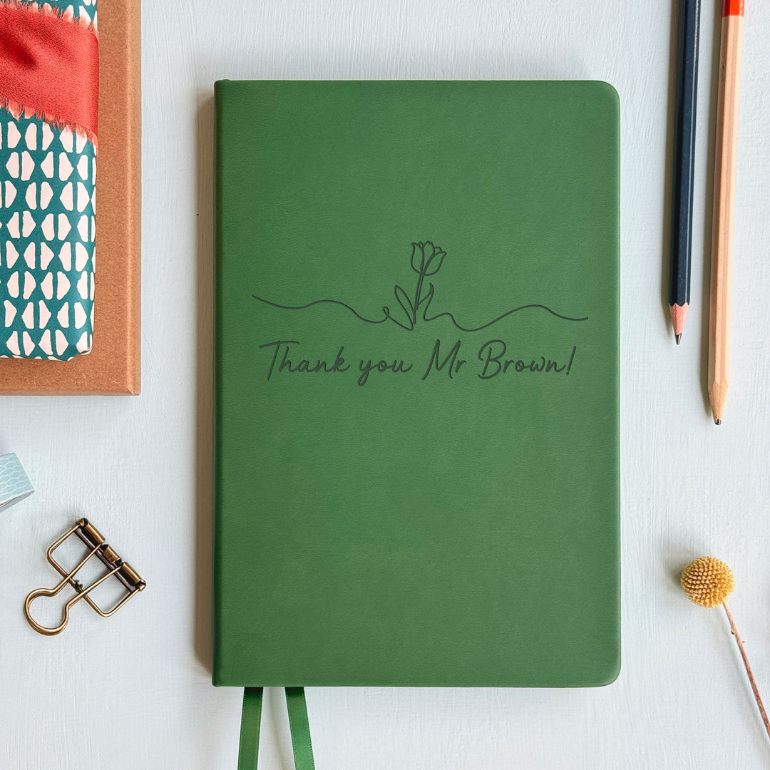 Floral motif tasteful end of year teacher gift forest green notebook on pale grey background surrounded by stationery and a dried flower.