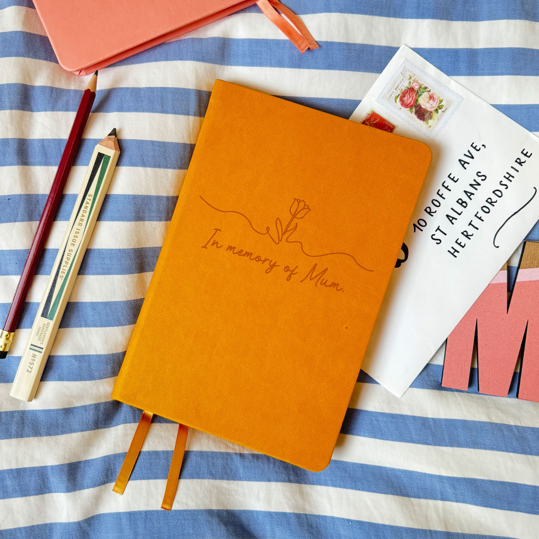 Floral motif remembrance funeral notes memorial notebook in memory of Mum engraved upon it. On a striped bedsheet atop a letter, surrounded by stationery items.