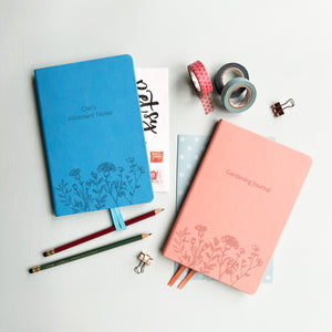 A top down image of two vegan leather notebooks, one blue and one pink. They are resting on a pale aqua table.  The fronts of the notebook feature a pattern of delicate flowers along the bottom edge. The personalised engraving on the blue notebook reads Dan's Allotment Notes and the personalised engraving on the pink notebook reads Gardening Journal. Surrounding the books are various stationary items.