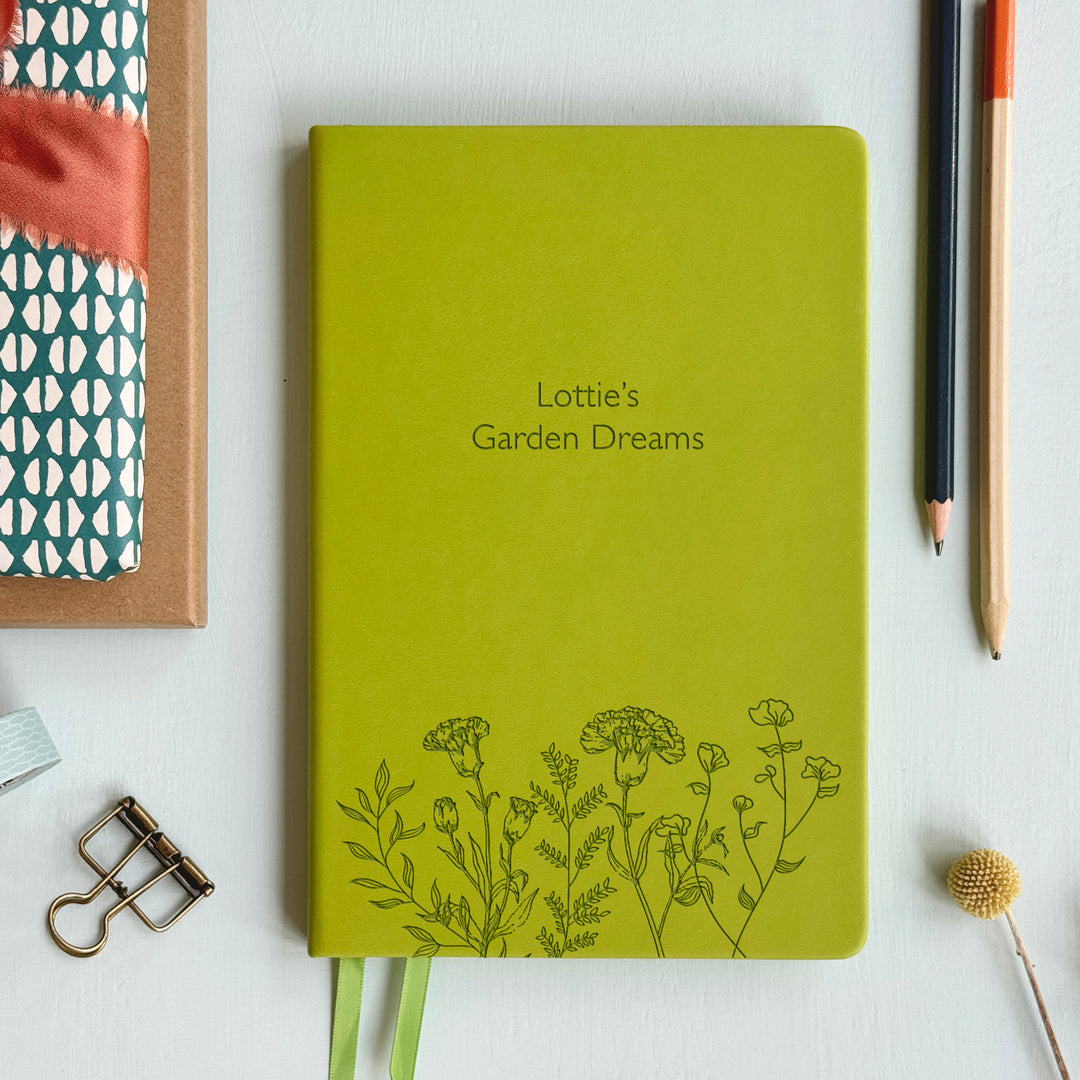 A top down image of a lime green vegan leather notebook resting on a pale aqua table. The front of the notebook features a pattern of delicate flowers along the bottom edge. The personalised engraving reads Lottie's Garden Dreams. Surrounding the book are various stationary items.