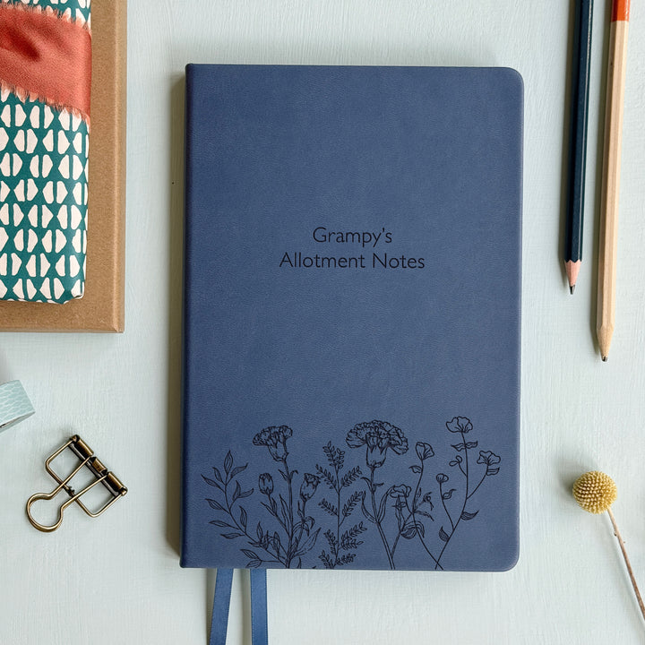 A top down image of a mid blue vegan leather notebook resting on a pale aqua table. The front of the notebook features a pattern of delicate flowers along the bottom edge. The personalised engraving reads Grampy's Allotment Notes. Surrounding the book are various stationary items.