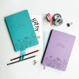 A top down image of two vegan leather notebooks, one turquoise and one lilac. They are resting on a pale aqua table.  The fronts of the notebook feature a pattern of delicate flowers along the bottom edge. The personalised engraving on the turquoise notebook reads Jack's Garden Dreams and the personalised engraving on the lilac notebook reads Nanny's Flower Notes. Surrounding the books are various stationary items.