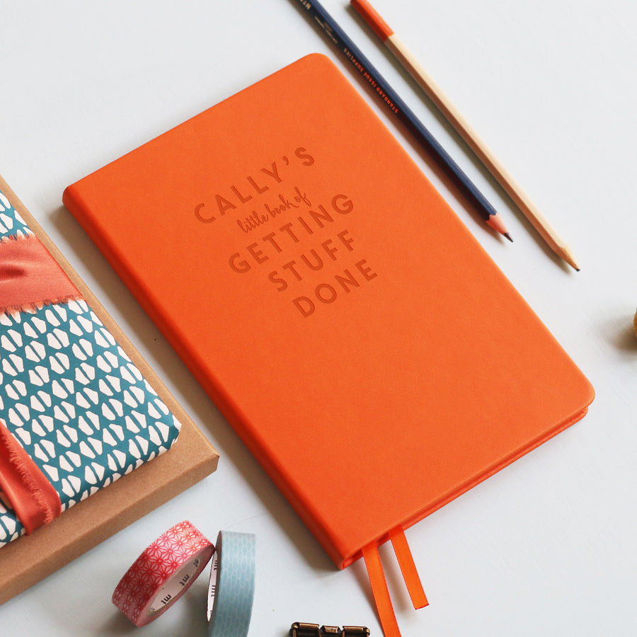 A top down image of a orange vegan leather notebook resting on a pale aqua table. The front of the notebook is personalised with the text  “Cally's little book of Getting Stuff Done". Surrounding the book are various stationary items.