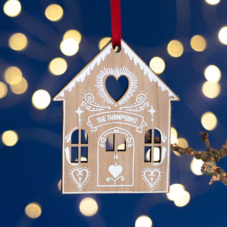 A wooden Christmas tree decoration shaped like a gingerbread house with white 'icing' and a red ribbon, personalized with the name 'Thompsons' and a heart shape cut out in the center.