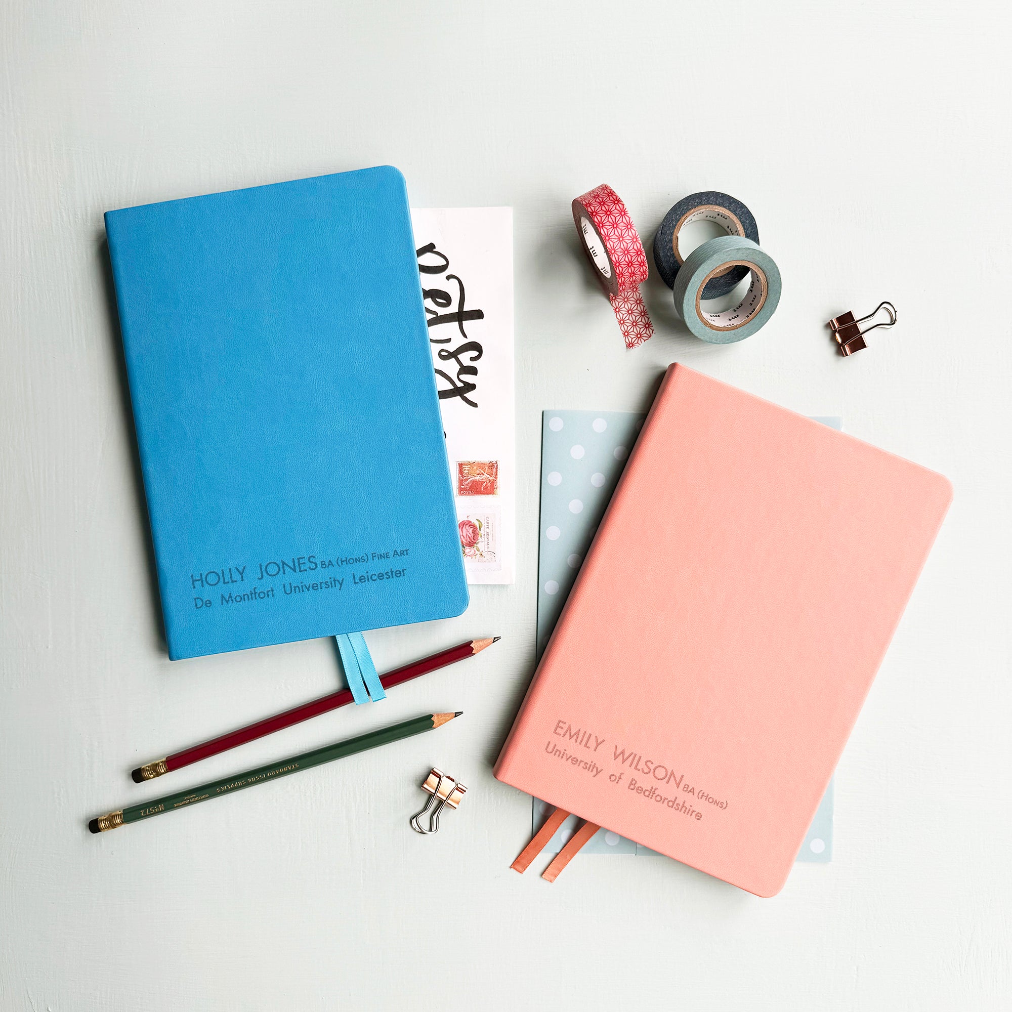 A top down image of two vegan leather notebooks, one blue and one pale pink. They are resting on a pale aqua table. The front of the blue notebook is engraved with the personalised text Holly Jones BA (hons) Fine Art, De Montfort University Leicester. The pink notebook reads Emily Wilson BA (hons), University of Bedfordshire. Surrounding the book are various stationary items.