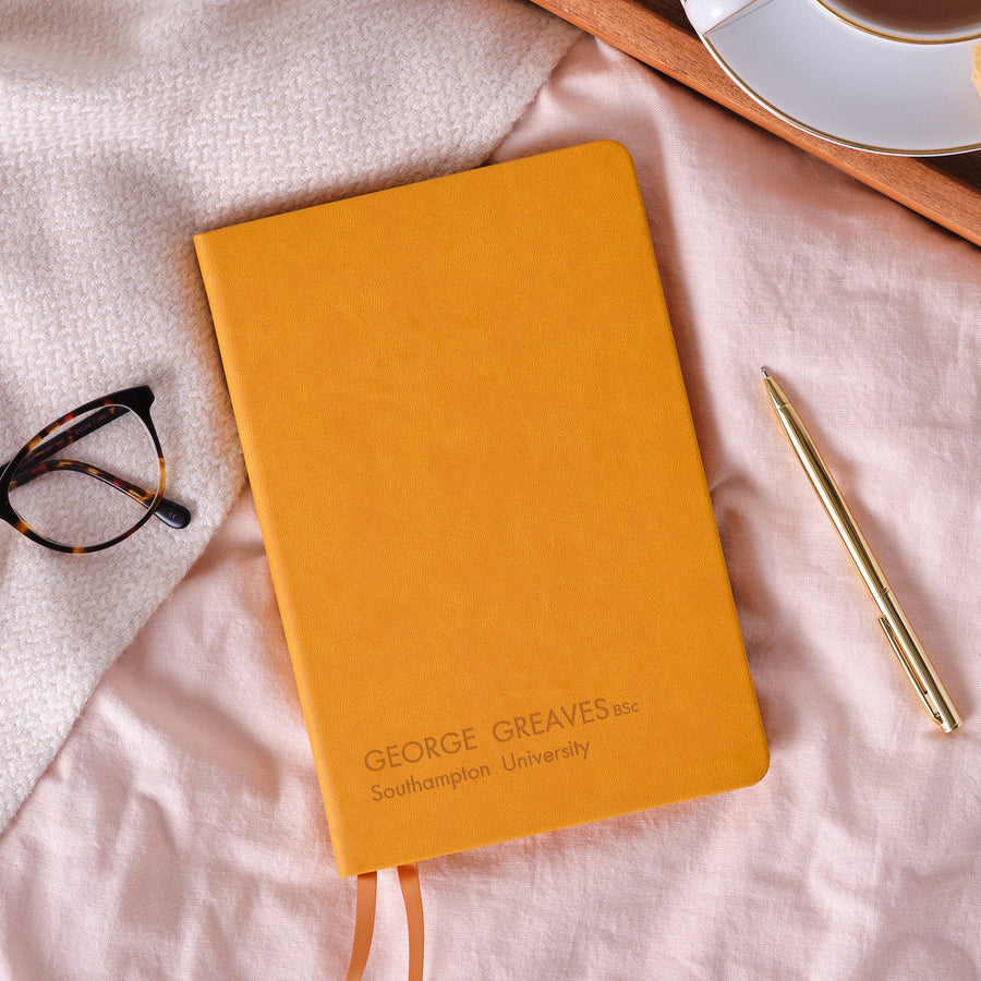 A top down image of a mustard yellow vegan leather notebook resting on a pale pink bed spread. The front of the notebook features personalised engraved text which reads - George Greaves BSc, Southampton University. Surrounding the book is a tray with a cup of tea and a biscuit, a pair of glasses and a gold pen.