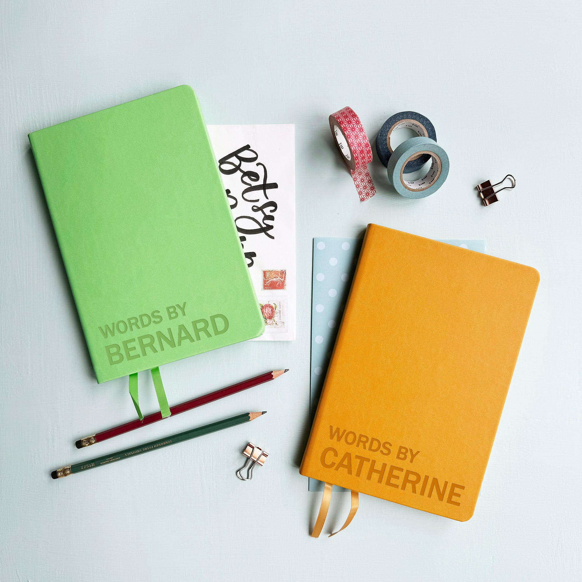 A top down image of two vegan leather notebooks, one green and one mustard yellow. They are resting on a pale aqua table. Both are engraved with the text Words By. The green notebook is personalised with the name Bernard and the mustard yellow book has the name Catherine.  Surrounding the book are various stationary items.