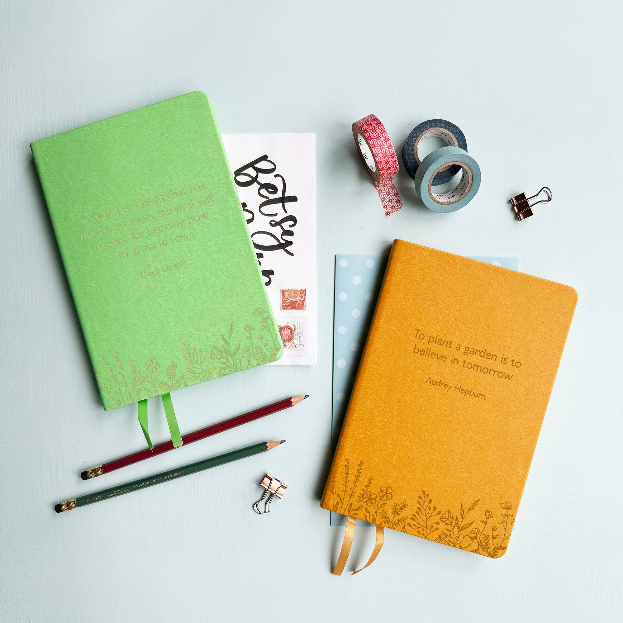 A top down image of two vegan leather notebooks, one green and one mustard yellow. The front of the notebook features a pattern of delicate flowers along the bottom edge. The personalised text has a quote from Doug Larson on the green notebook and on the mustard yellow a quote from Audrey Hepburn.  Surrounding the book are various stationary items.