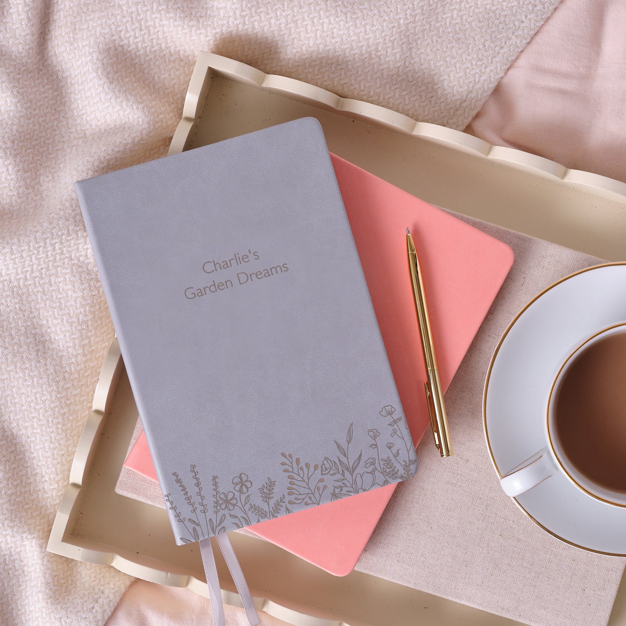 A top down image of a grey vegan leather notebook. The front of the notebook features a pattern of delicate flowers along the bottom edge.  The personalised engraving reads “Charlie’s Garden Dreams”. It is stacked above a pink notebook and is resting on a cream wooden tray with a cup of tea, and a gold pen beside it. The tray is resting on a pink bedspread.