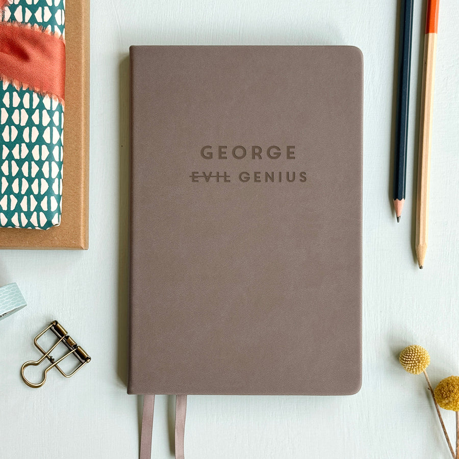 A top down image of a grey vegan leather resting on a pale aqua table. The front of the notebook is personalised with the name George and engraved with the phrase Evil Genius. Surrounding the book are various stationary items.