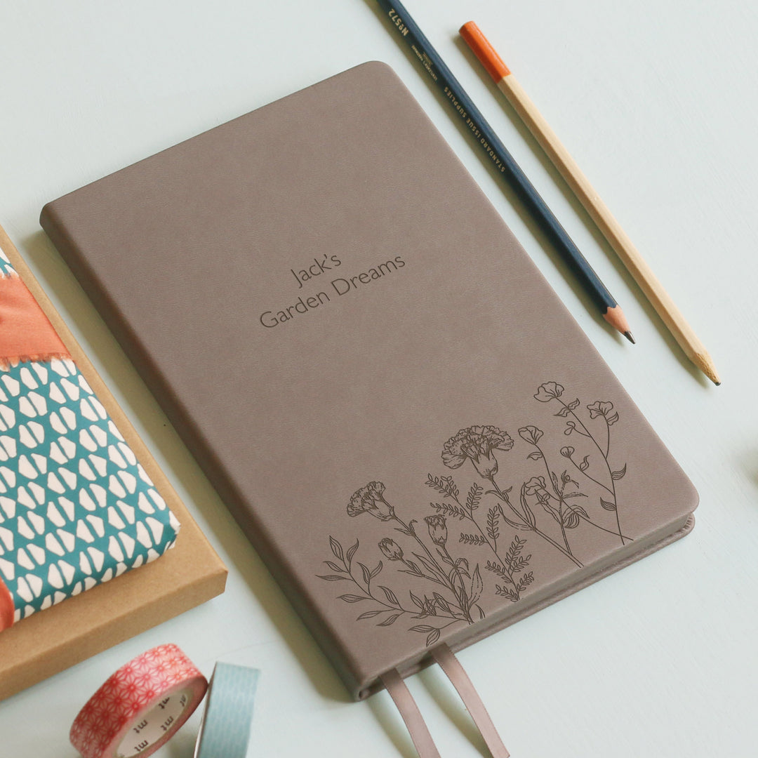 A top down image of a grey vegan leather notebook resting on a pale aqua table. The front of the notebook features a pattern of delicate flowers along the bottom edge. The personalised engraving reads Jack's Garden Dreams. Surrounding the book are various stationary items.