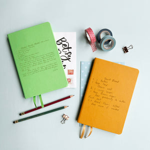 A top down image of two vegan leather notebooks, one green and one mustard yellow. They are resting on a pale aqua table. Both books are engraved with a handwritten recipe.  Surrounding the book are various stationary items.