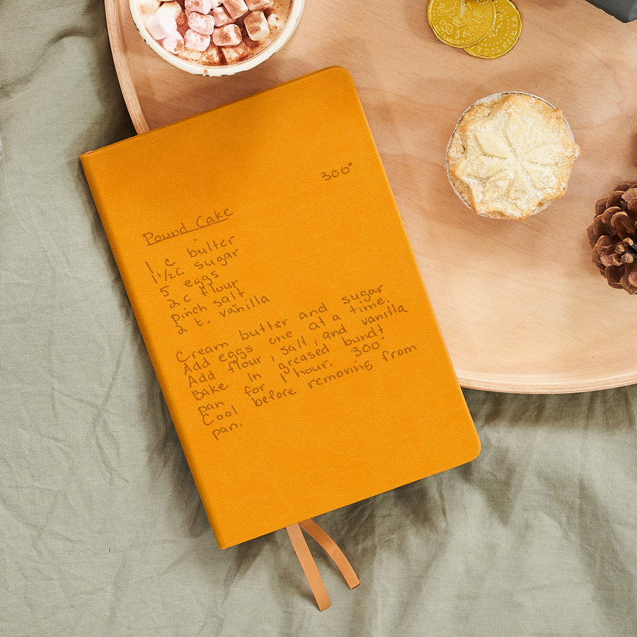A top down image of a mustard yellow vegan leather notebook. The book is engraved with a handwritten recipe. It is resting on a wooden tray with a mug of hot chocolate, a mince pie, and a pine cone.