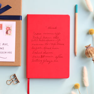 A top down image of a red vegan leather notebook resting on a pale aqua table. The book is engraved with a handwritten recipe. Surrounding the book are various stationary items and dried flowers.