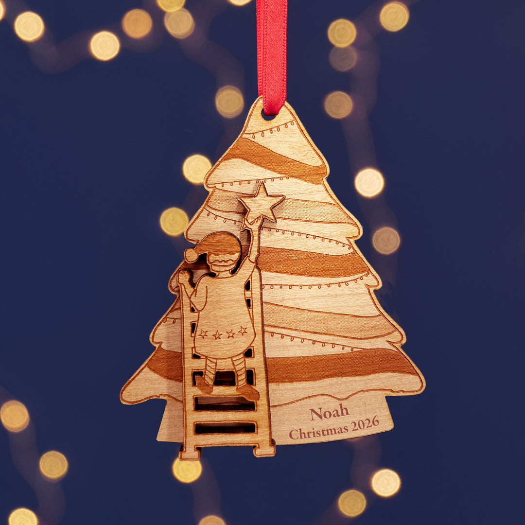 Personalised wooden Christmas tree decoration with a child climbing a ladder to put the star on the of the tree. The text reads Noah Christmas 2026. The background is dark blue with fairy lights.